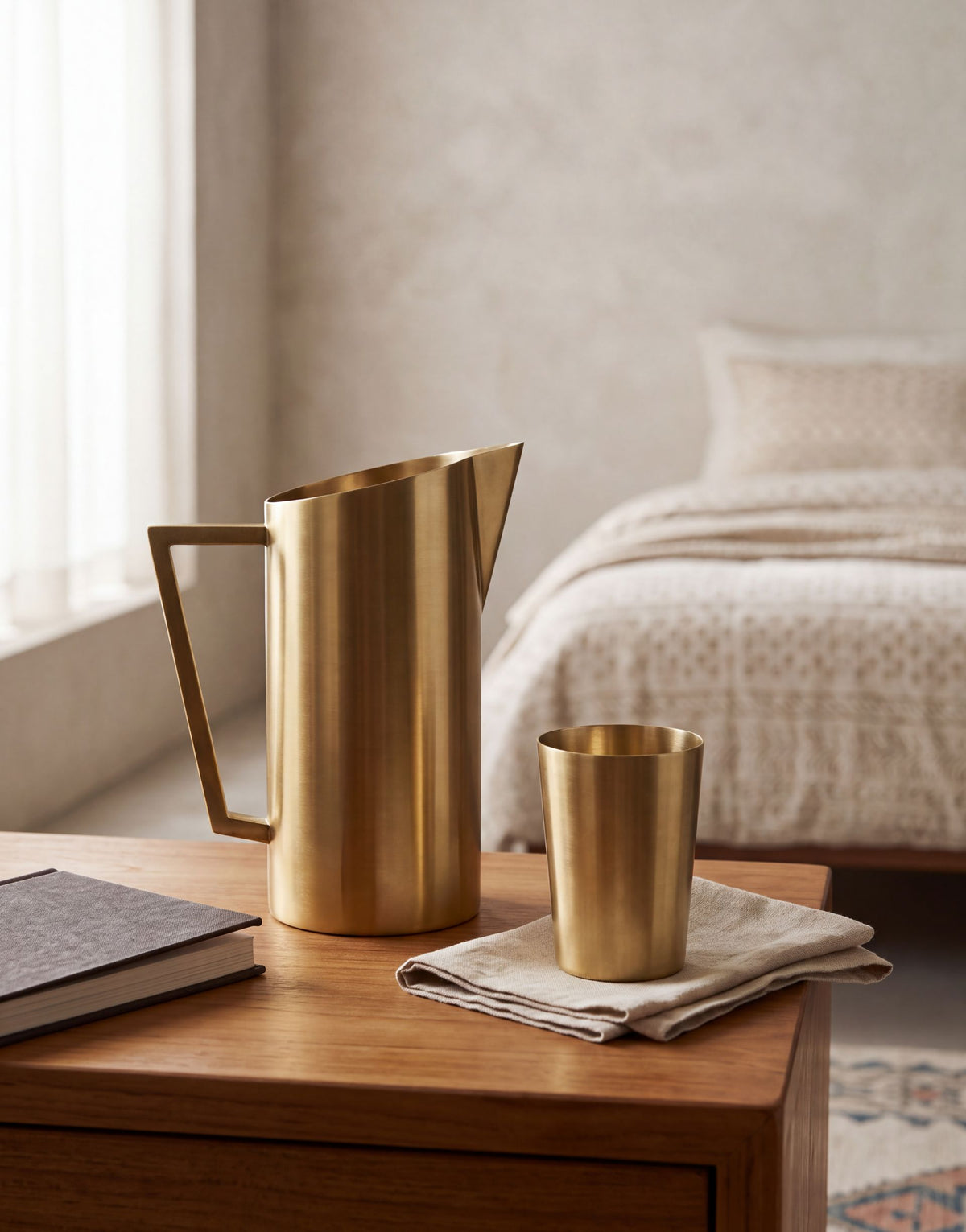 A brushed brass pitcher and matching cup set on a wooden bedside table in a softly lit bedroom. The cup rests on a folded linen napkin, with a bed and neutral wall visible in the background.