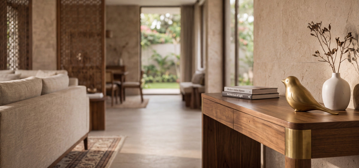 A warm, modern interior scene with a wooden console table in the foreground holding a brass bird figurine, books, and a white vase with dried branches. A beige sofa is visible on the left, and an open doorway in the background reveals a garden view.