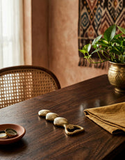 Wooden table with brass bottle opener and a plant in a room with a textured wall and woven chair.