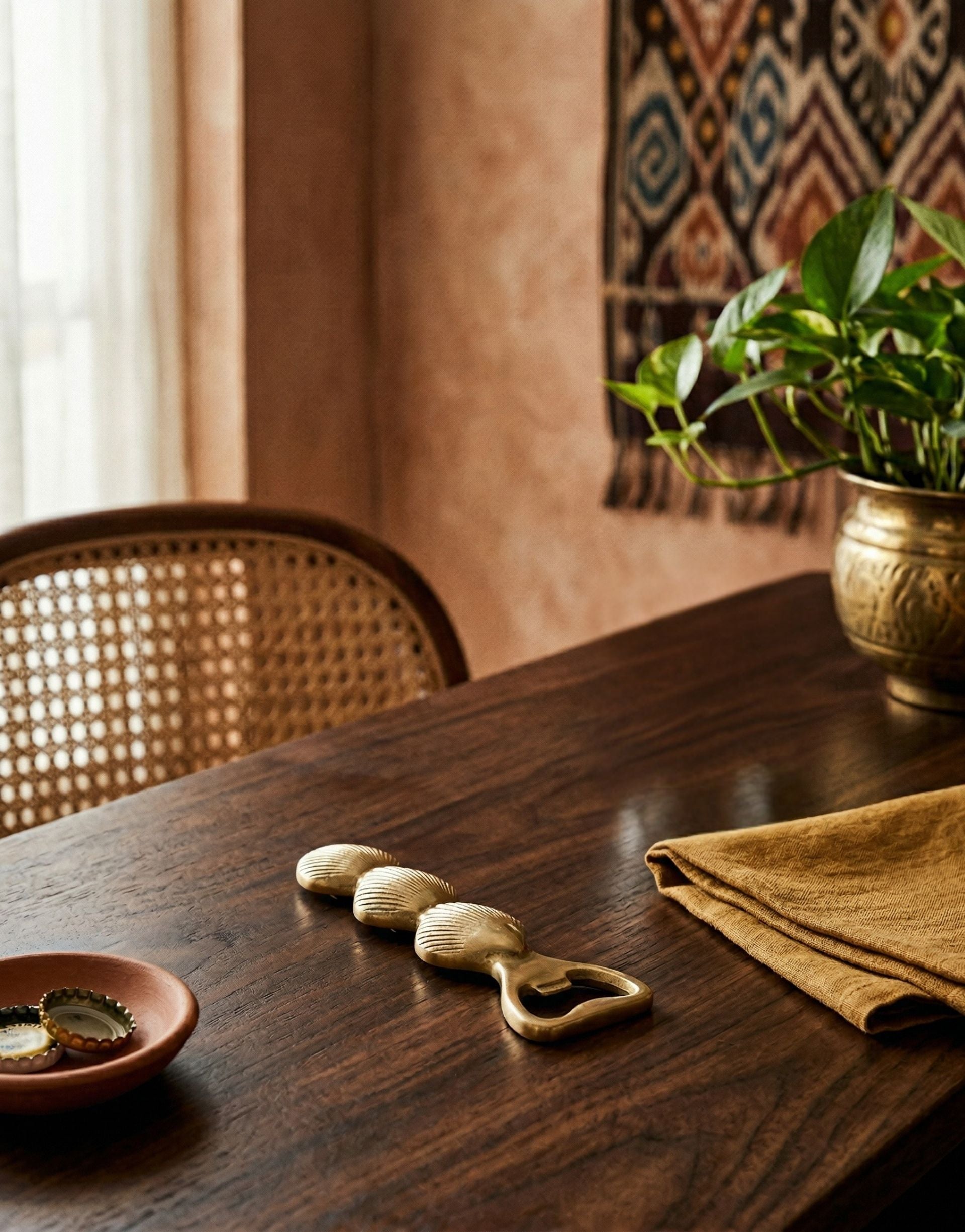 Wooden table with brass bottle opener and a plant in a room with a textured wall and woven chair.