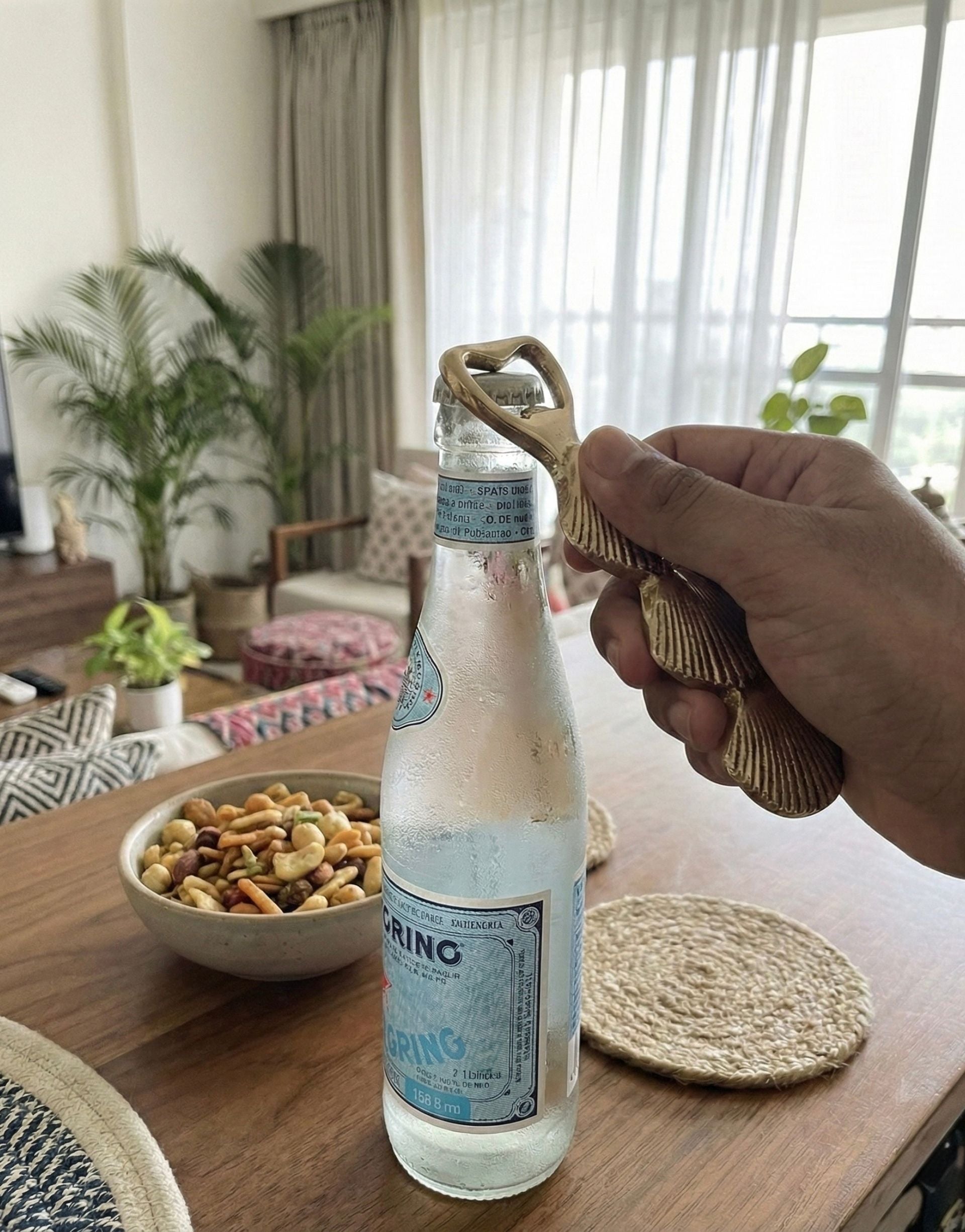 Hand holding a bottle opener on a wooden table with snacks and a living room background