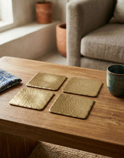 Set of four brass coasters on a wooden table with a cup and folded fabric in a cozy room.
