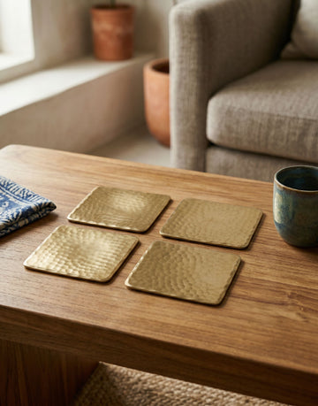 Set of four brass coasters on a wooden table with a cup and folded fabric in a cozy room.