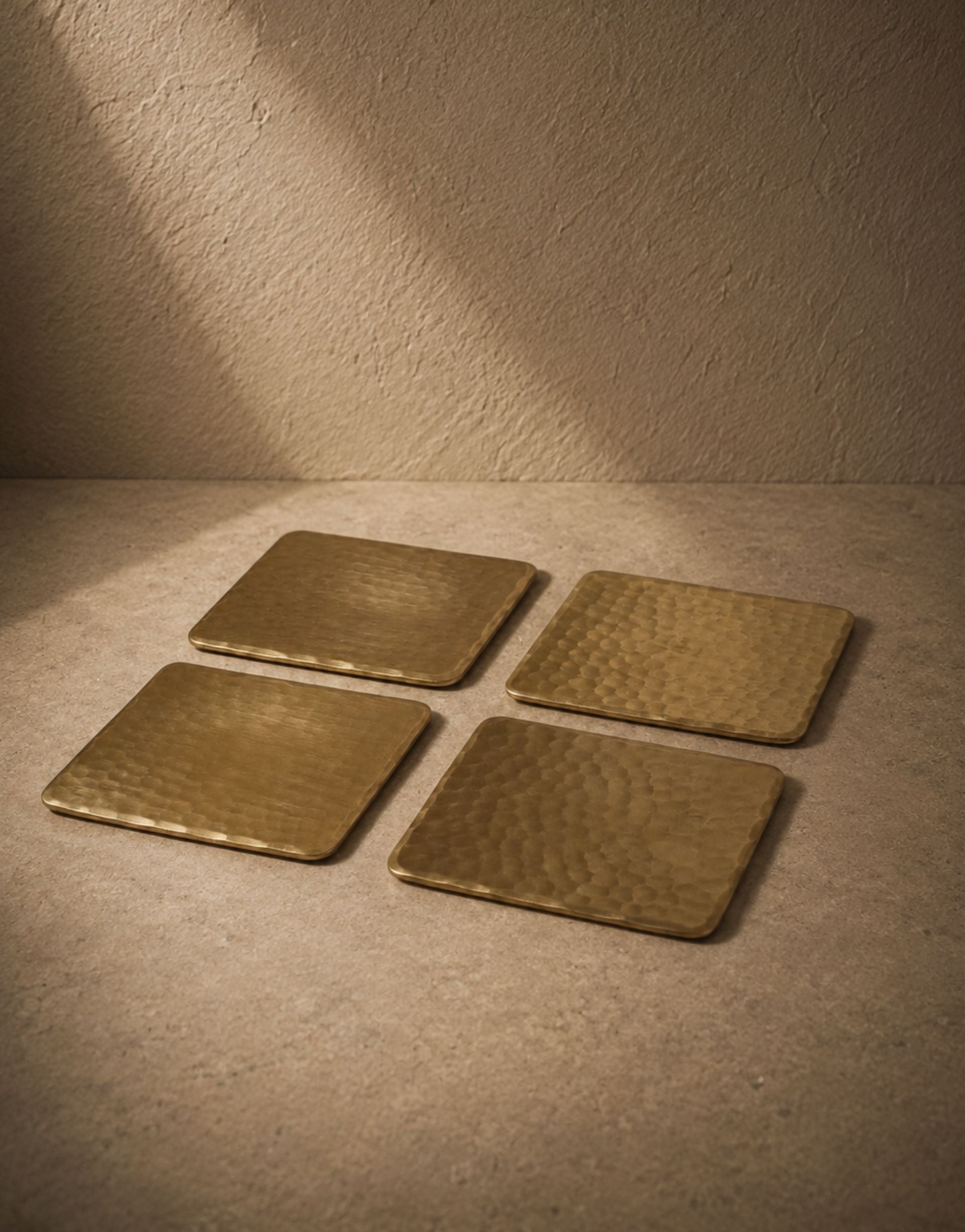 Four square brass coasters on a textured beige surface