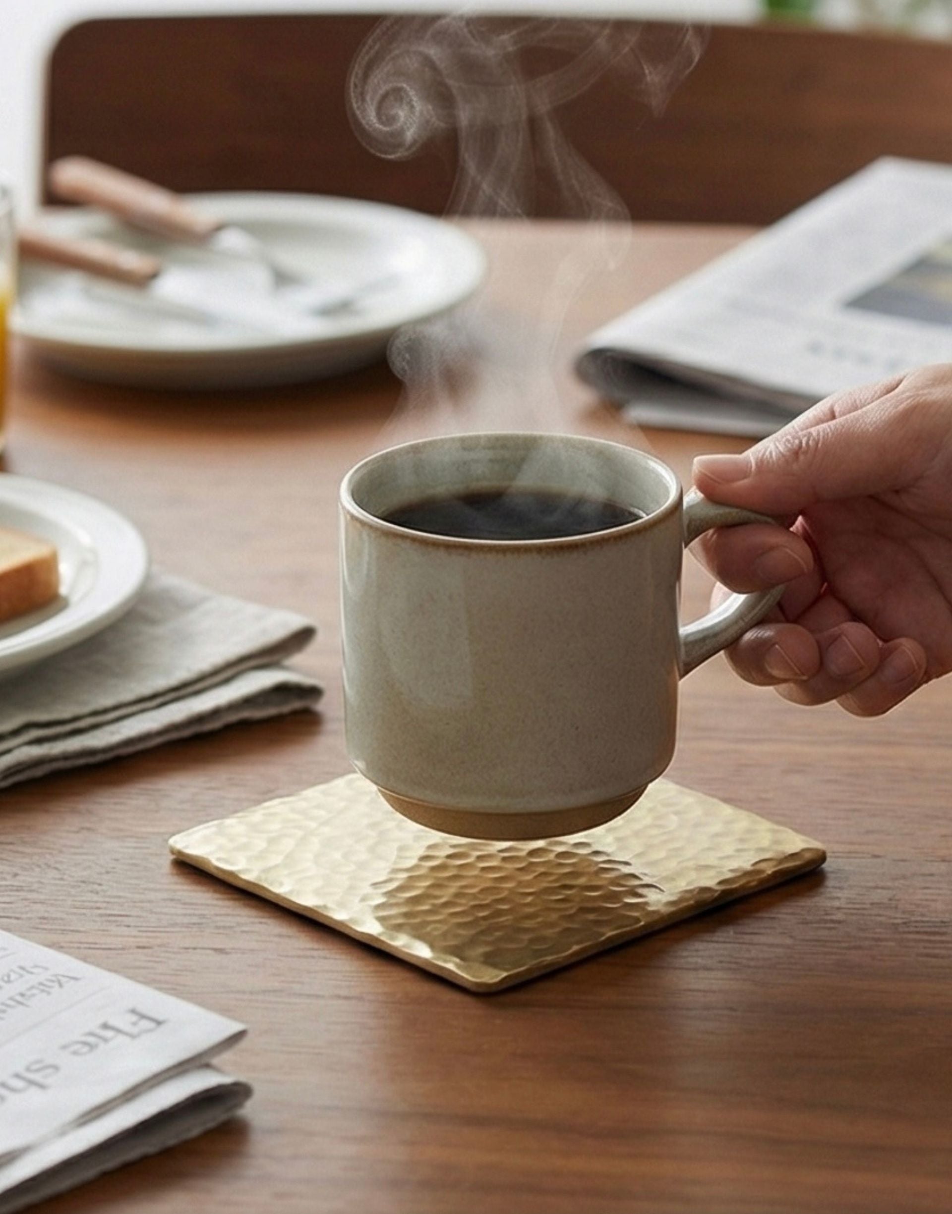 Person holding a steaming mug of coffee on a brass coaster on a wooden table with plates and a newspaper.