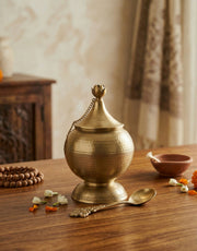 Brass ghee container with a chain on a wooden table with decorative elements.
