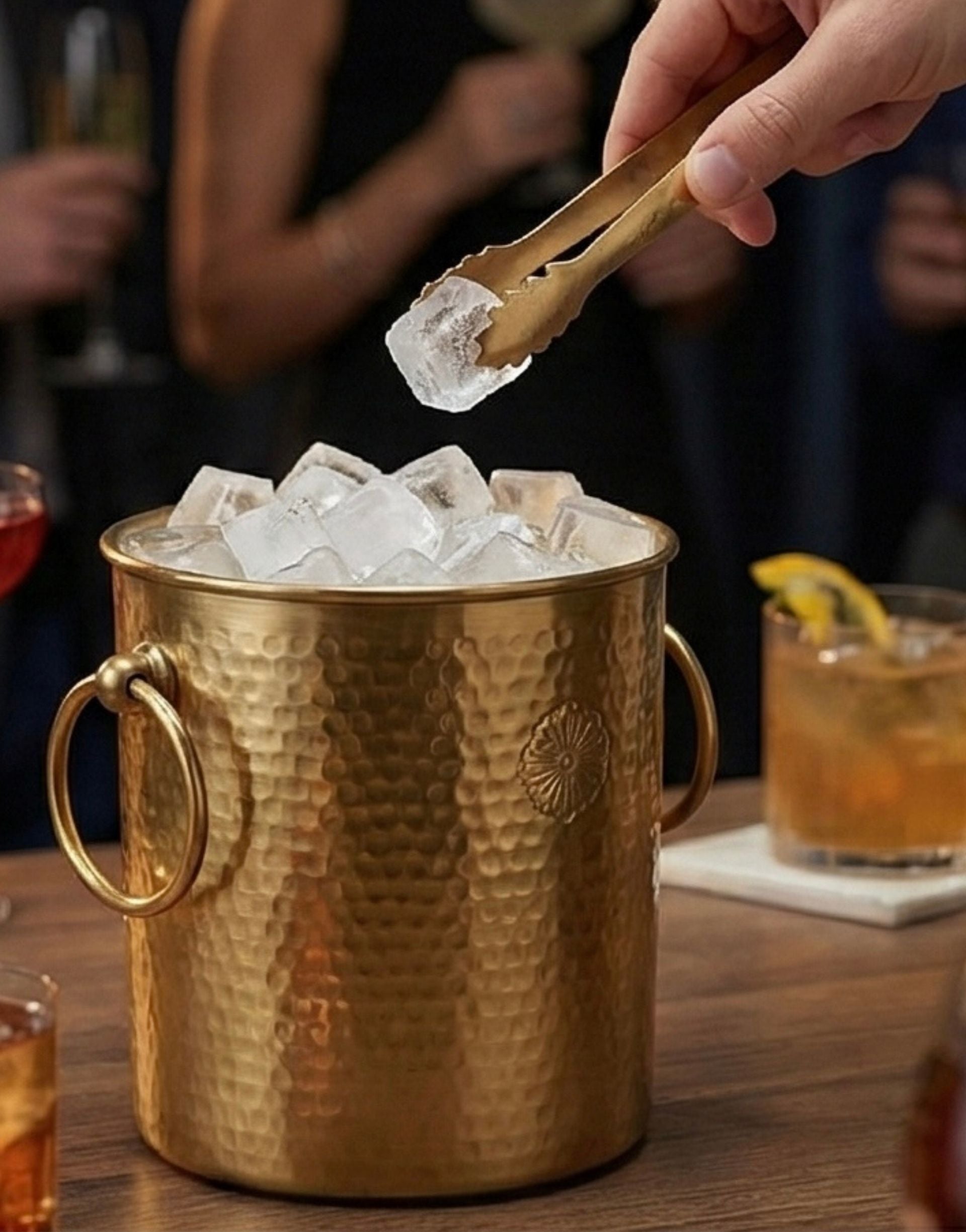 Brass ice bucket with ice cubes and a hand using tongs, with a blurred bar setting in the background.