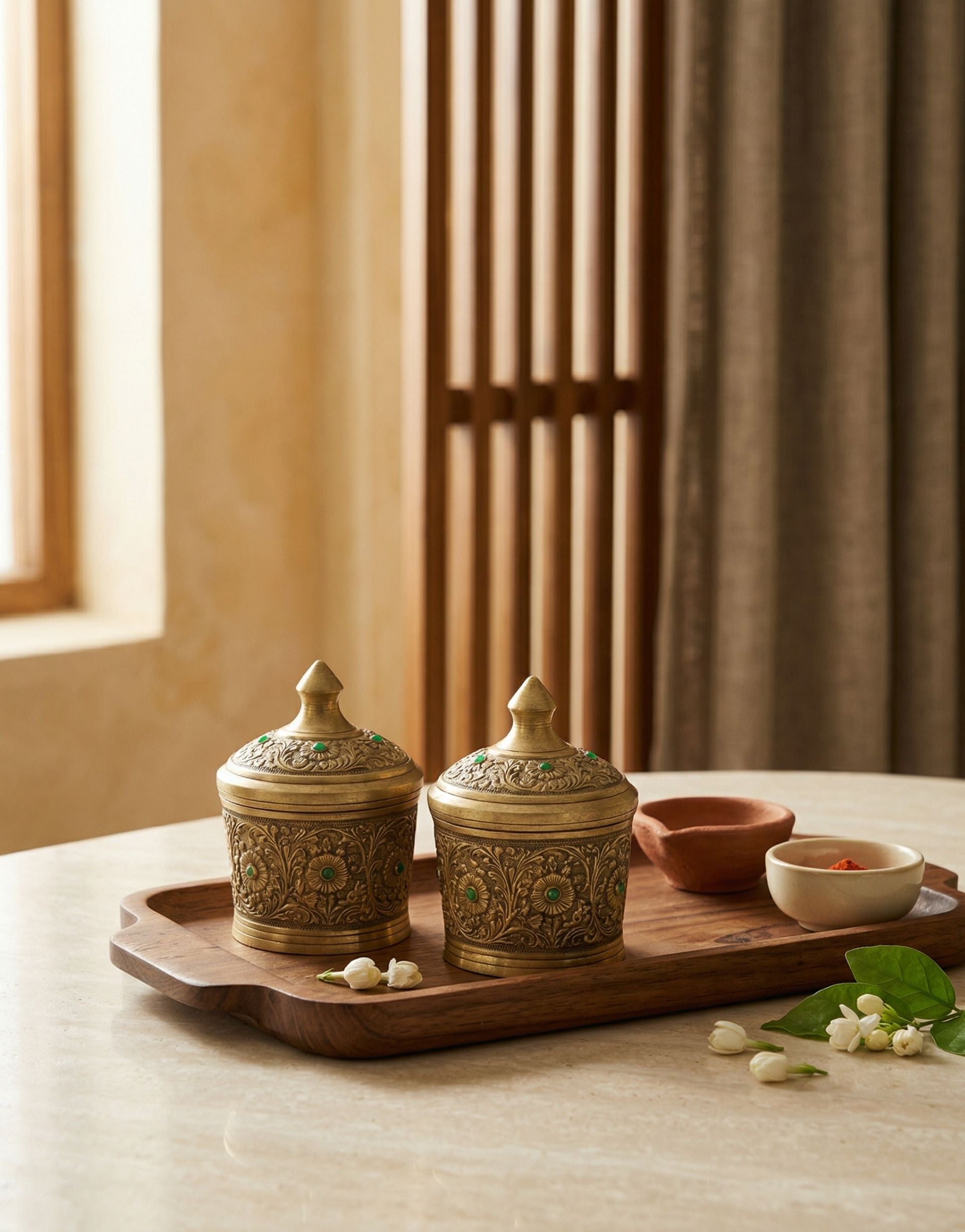 Decorative brass kumkum and rice containers with intricate designs on a wooden tray, set against a warm-toned curtain.