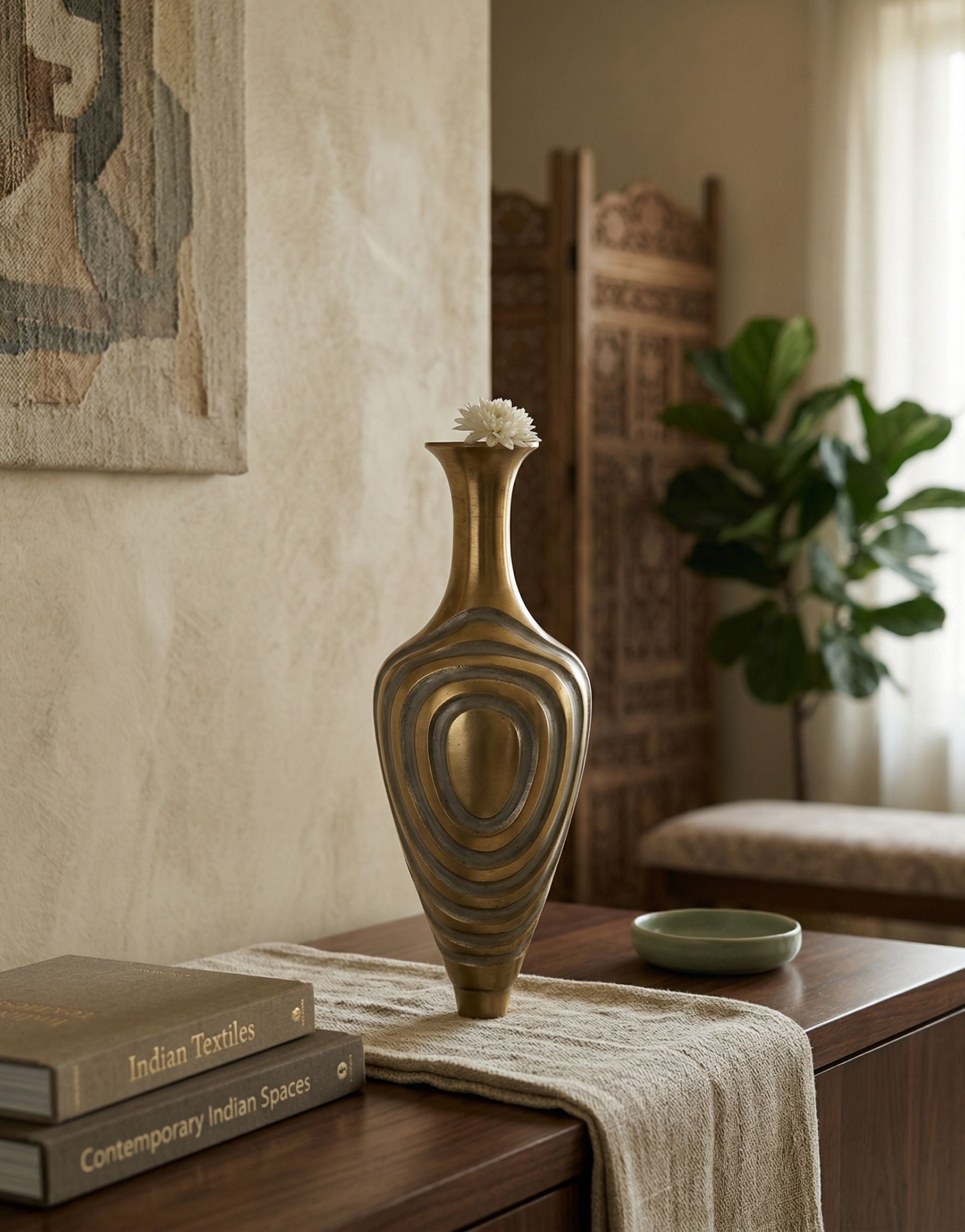 Decorative brass vase on a table with books and a plant in the background