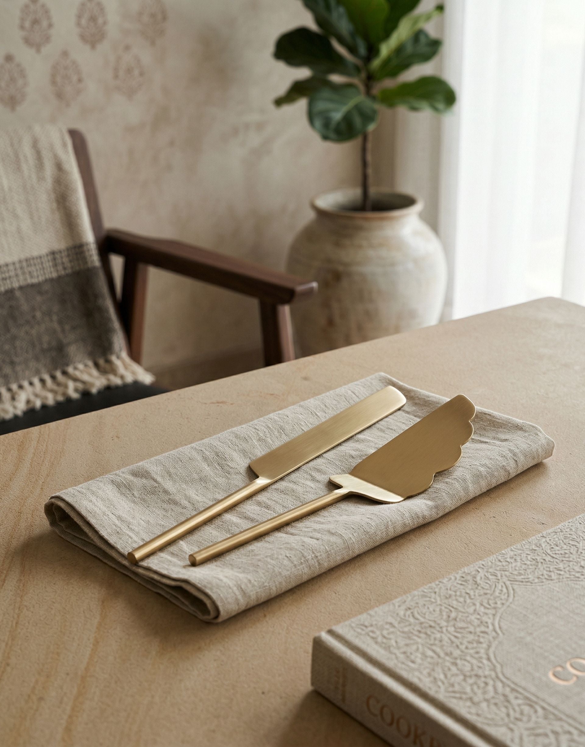Brass cake knife and server set on a beige napkin with a plant and chair in the background