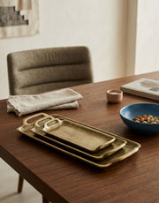 Brass trays on a wooden table with a bowl of nuts and a chair in the background.