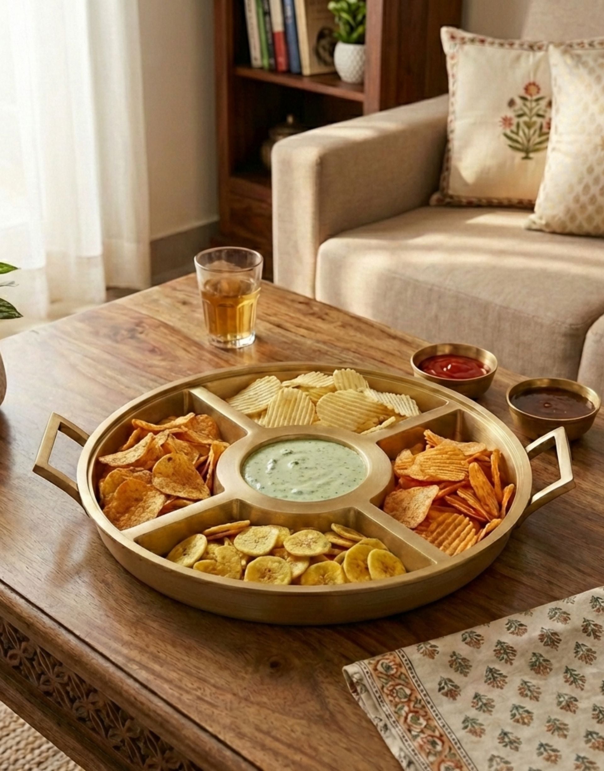 Round brass snack platter with compartments filled with chips and dips on a wooden table in a cozy living room.