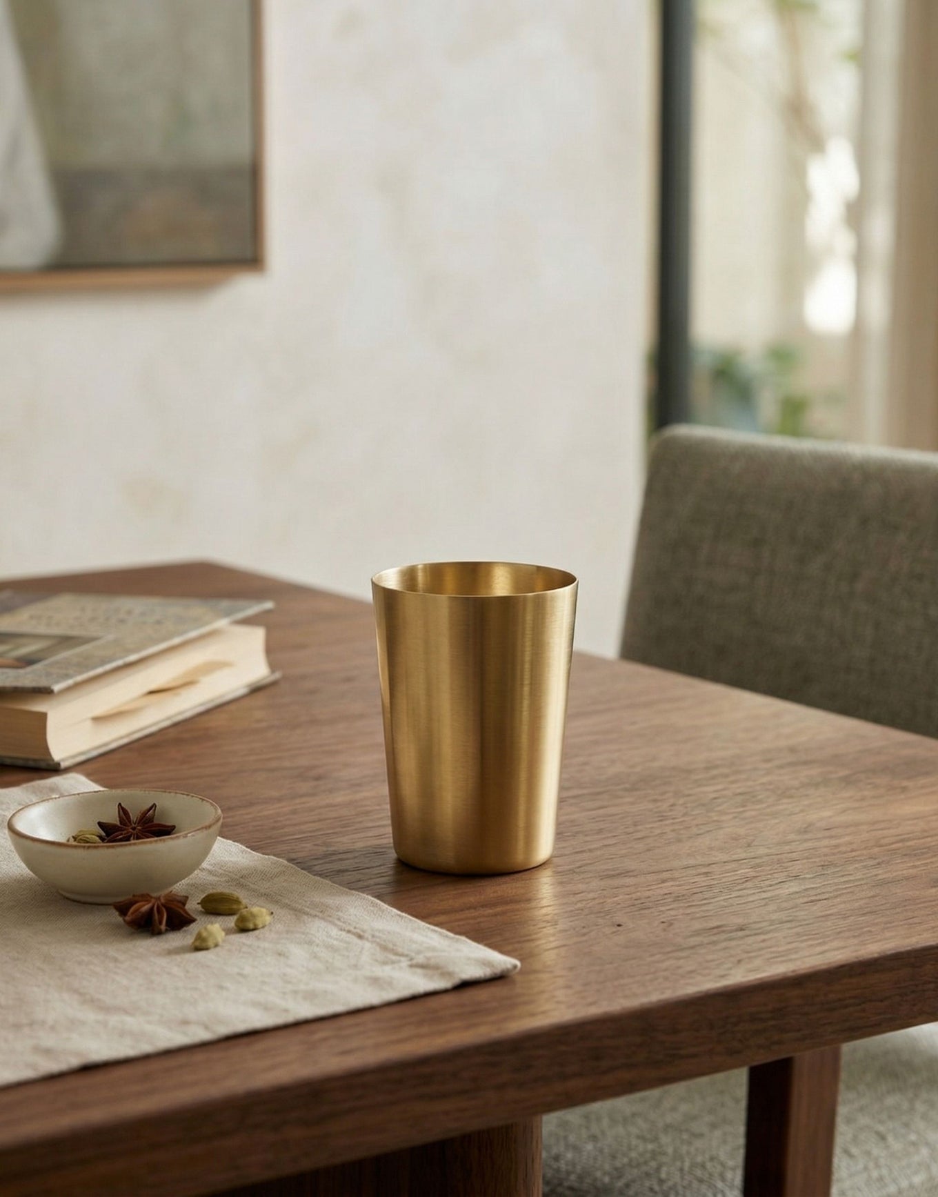 Brass tumbler on a wooden table with a book and small bowl in a softly lit room.