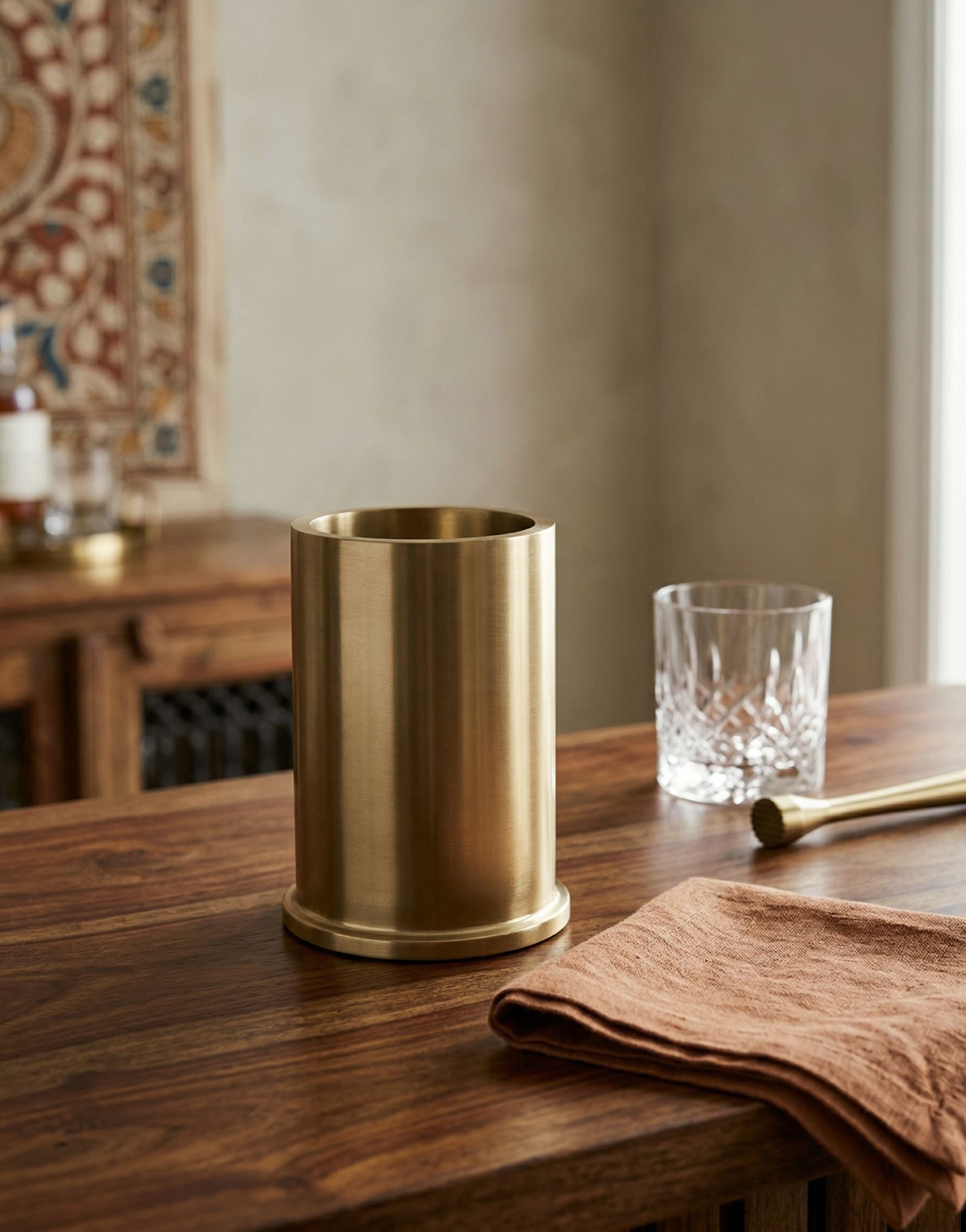 Brass wine chiller on a wooden table with a glass and napkin in a home setting.