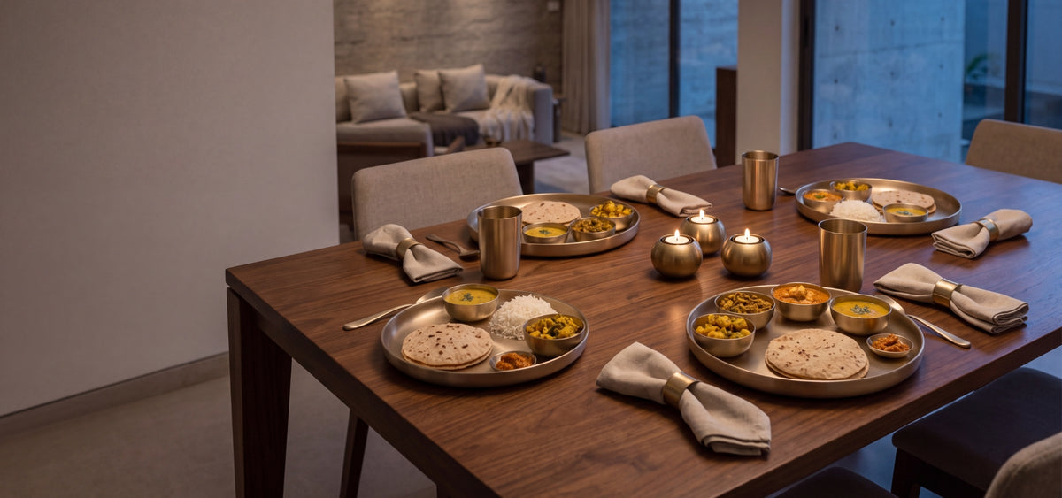 A wooden dining table set for a meal with kansa thali of Indian food, small brass bowls, folded beige napkins in brass table napkin rings, and lit candles  in a brass candle holderin a modern dining room.