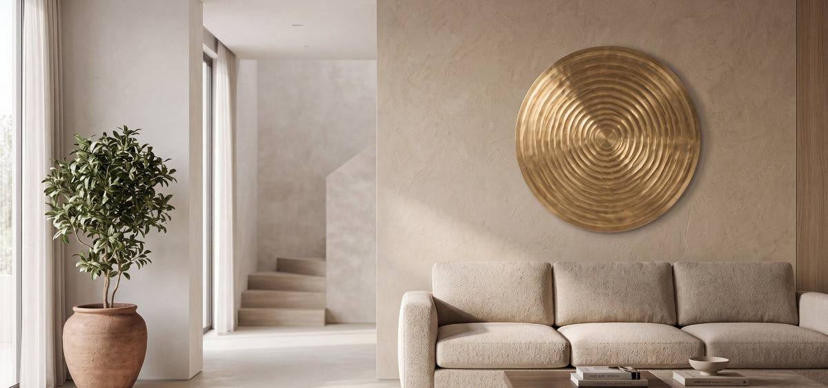 A large circular brass wall art piece with concentric rings is mounted on a textured beige wall above a light beige sofa in a modern, minimalist living room.
